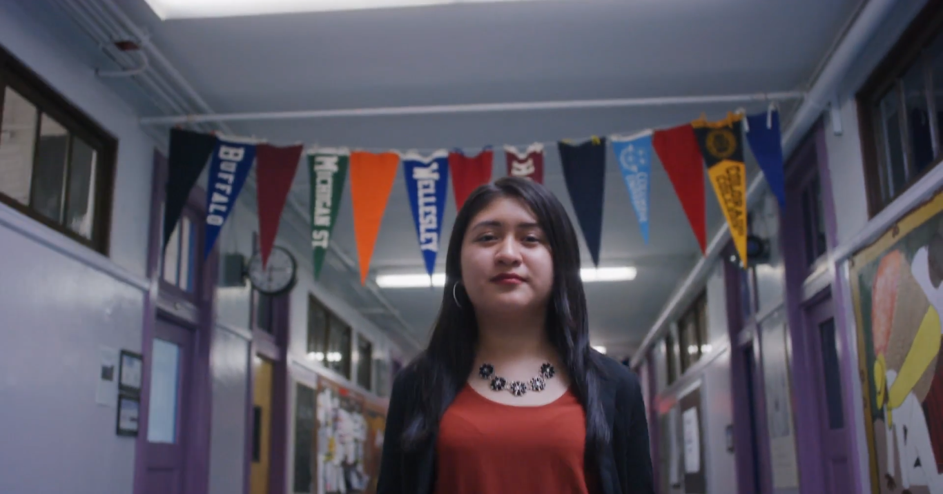 Cinthia Ibarra walks through a school hallway with college banners hanging behind her.
