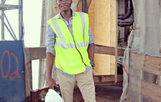 Gideon stands at a construction site in a yellow safety vest, holding a construction helmet in his right hand.